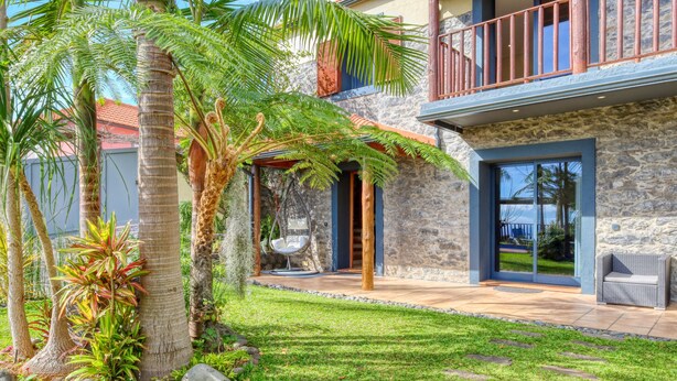 A modern house with a stone facade, a balcony with a wooden railing, and a well-maintained garden with palm trees and a lawn. - Ricardo House, a Home in Madeira (Calheta)