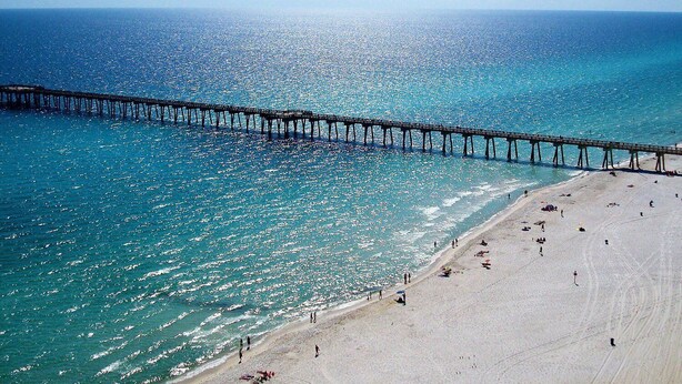 A beach with a pier extending into the sea, sandy shore, and clear blue water.