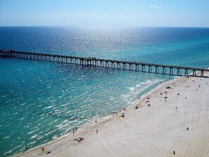 A beach with a pier extending into the sea, sandy shore, and clear blue water.