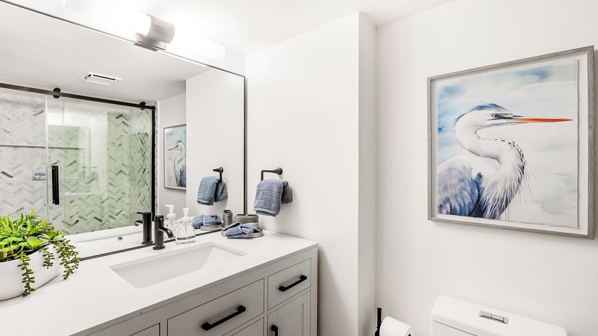 A modern bathroom with a white vanity, a large mirror, a framed painting of a bird, and a potted plant.