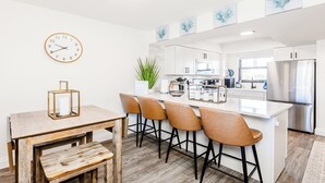 Une cuisine moderne avec des meubles blancs, un îlot central doté de tabourets, une horloge au mur et une plante en pot.