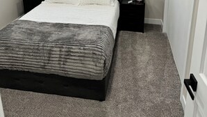 A bedroom with a bed, two bedside tables, a window with blinds, and a door.