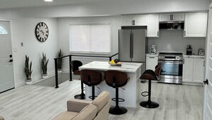 A modern kitchen with white cabinets, a stainless steel refrigerator, and a built-in oven. There is a dining area with a white island and bar stools.