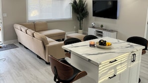 A modern living room with a sofa, a dining area with a marble table, and a TV mounted on the wall.