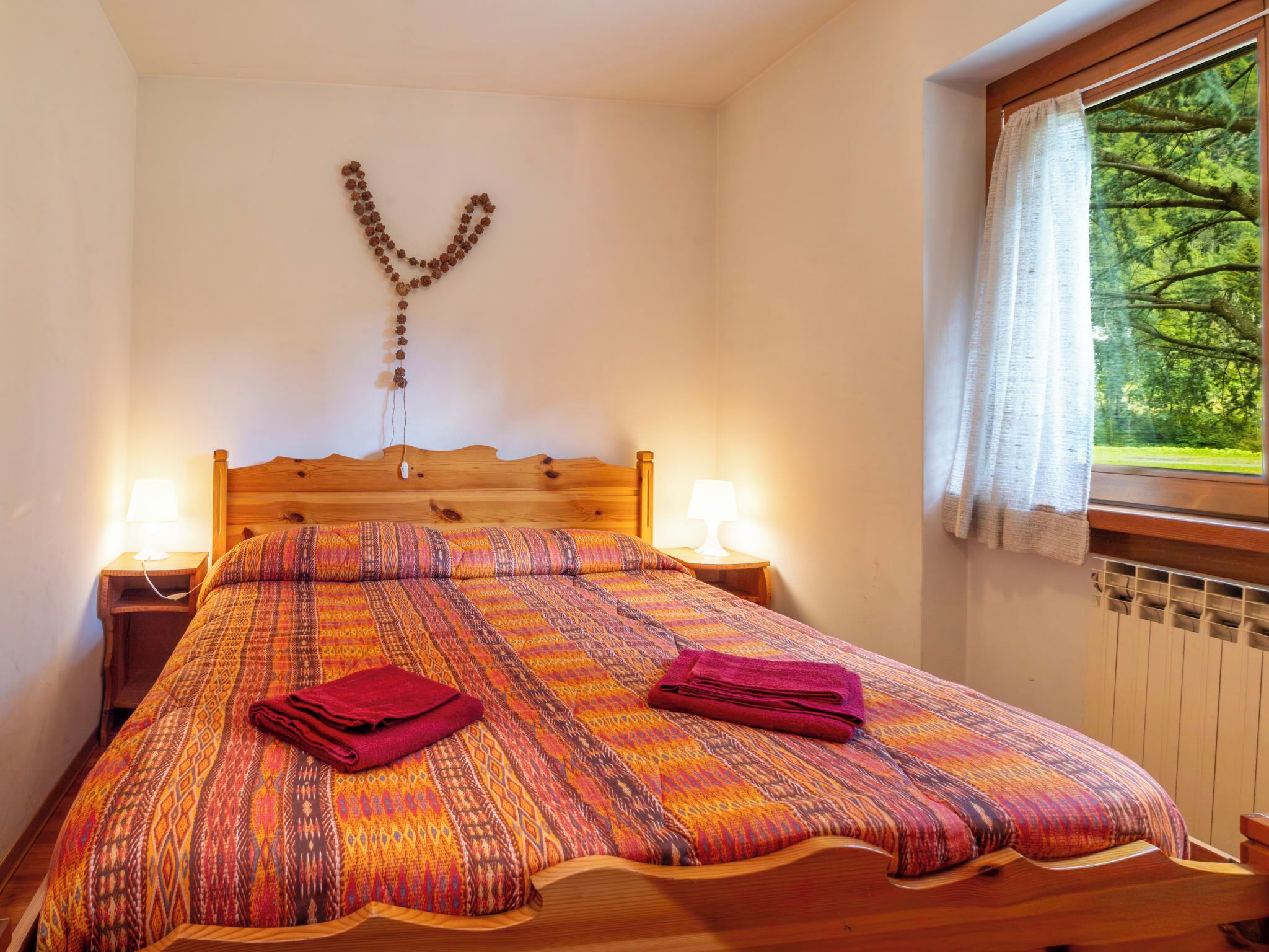 Ein Schlafzimmer mit einem hölzernen Bett, gemusterten Betttextilien, zwei Nachttischlampen, einer Wanddekoration und einem Fenster mit Blick auf Bäume.