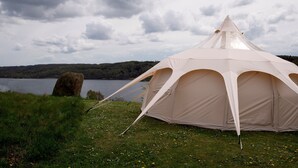 A large, beige tent with a conical roof, set on a grassy area near a lake with a forested hill in the background. - Bramslev Bakker Naturhotel (Hobro)