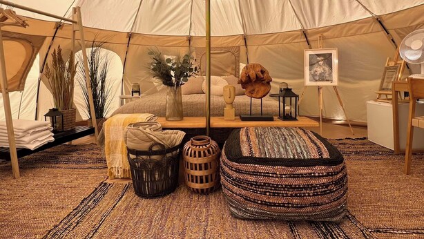 A cozy tent interior with a bed, a small table, a hanging plant, and a woven pouf. - Bramslev Bakker Naturhotel (Hobro)