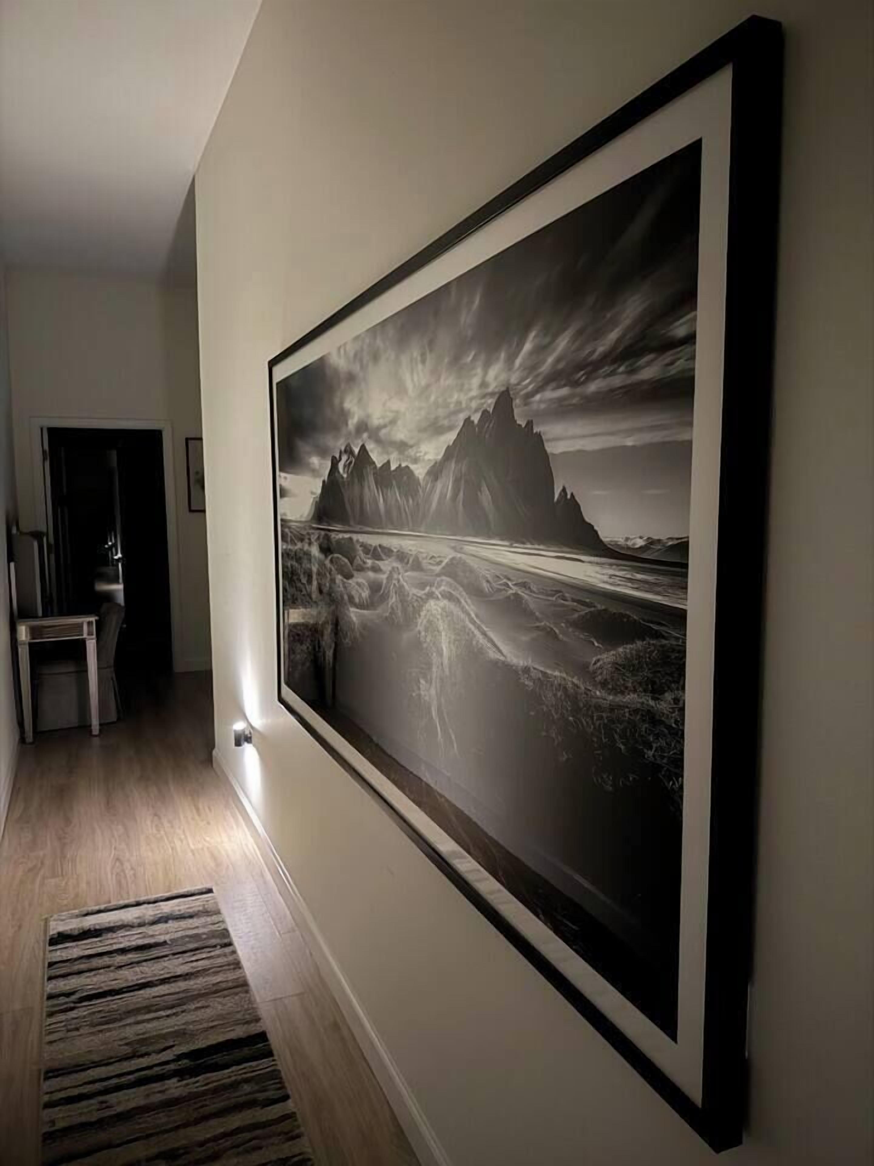 Een gang met een zwart-wit landschapsfoto in een lijst, een houten vloer en een vloerkleed.