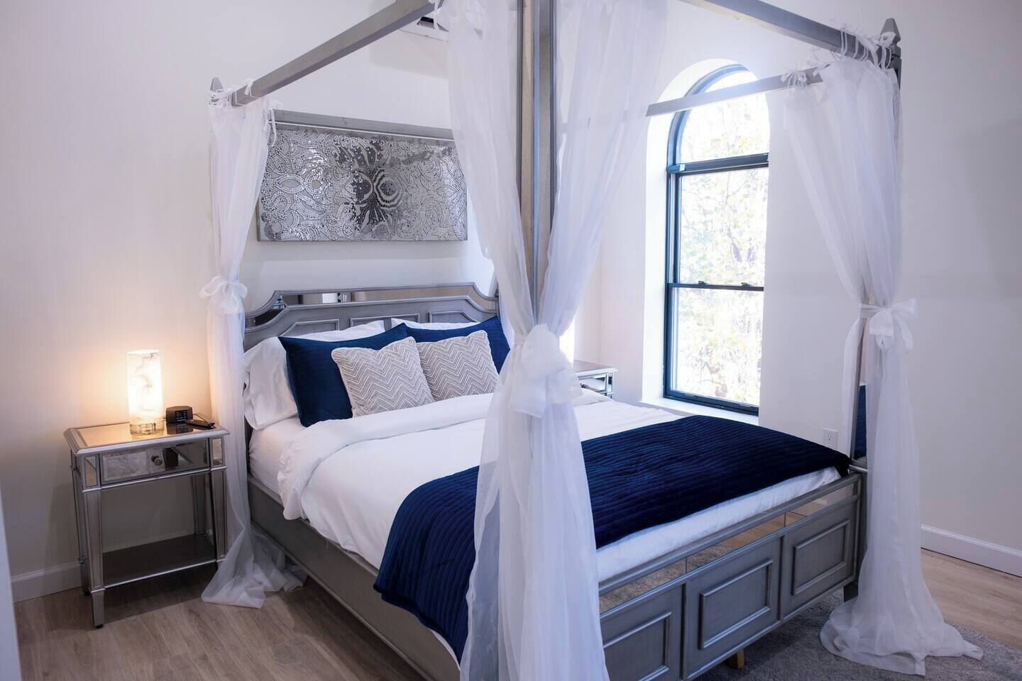 Een hemelbed met nachtkastje, bedlamp en raam met uitzicht.
