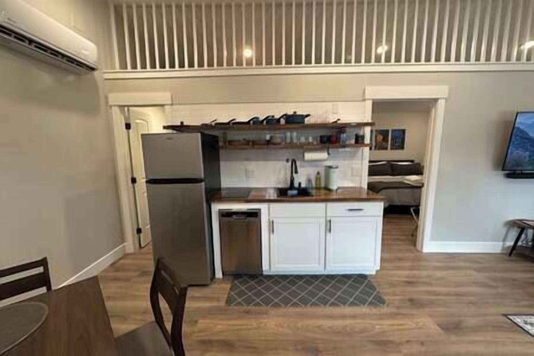 Una cocina compacta equipada con refrigerador, lavaplatos y pileta. Cuenta con un pequeño comedor con mesa y sillas. El diseño de la cocina es moderno, con alacenas blancas y una encimera de madera.