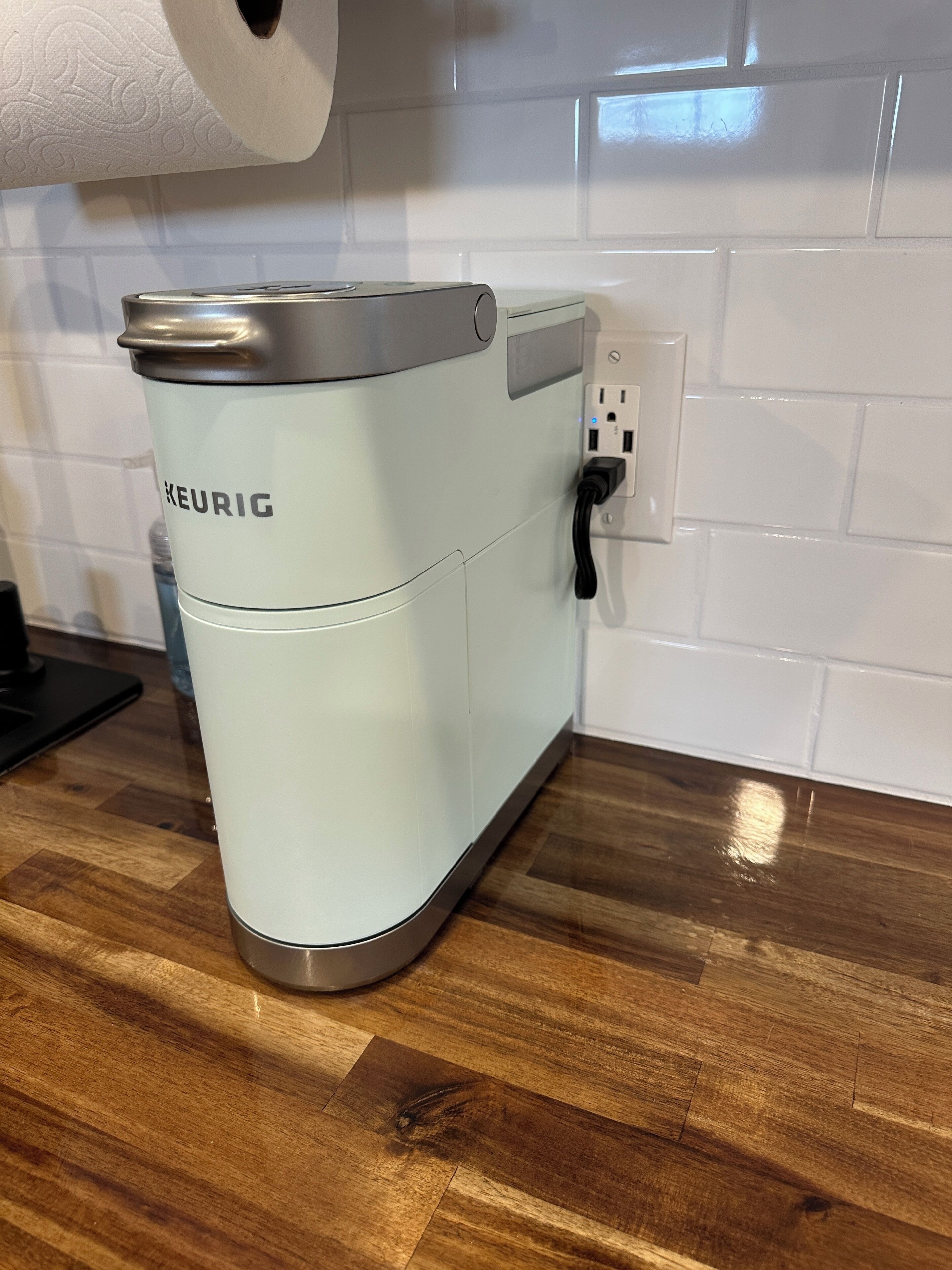 Une machine à café Keurig posée sur un comptoir en bois, avec un dosseret de carreaux blancs.