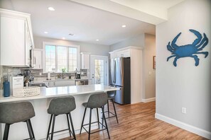 A modern kitchen with white cabinetry, a large island with bar stools, a stainless steel refrigerator, and a blue crab wall decoration.