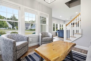 A spacious living room with large windows, a wooden coffee table, two patterned armchairs, and a staircase with white railings.