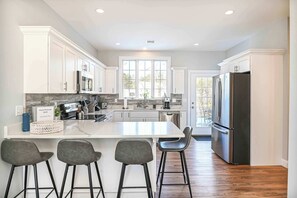 A modern kitchen with white cabinets, a large island with bar stools, stainless steel appliances, and a view to the outside.
