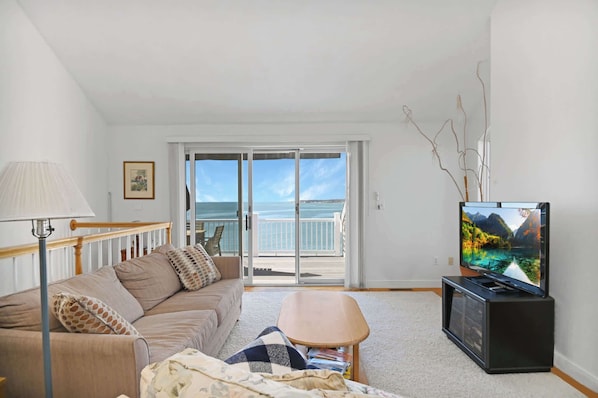 A living room with a sofa, a coffee table, a TV on a stand, a lamp, and a view of the sea through a glass door.