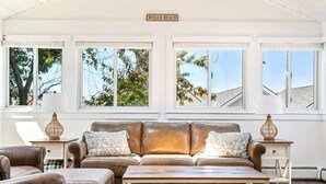 A living room with a brown leather sofa, a wooden coffee table, and two side tables with lamps. There are three windows with a view of trees and a building. - Near Crescent + Wells Beach • Firepit • Sleeps 10 (Wells)