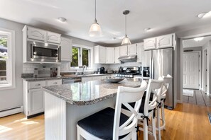 Una cocina moderna con armarios blancos, electrodomésticos de acero inoxidable, una isla con encimera de granito e iluminación colgante.