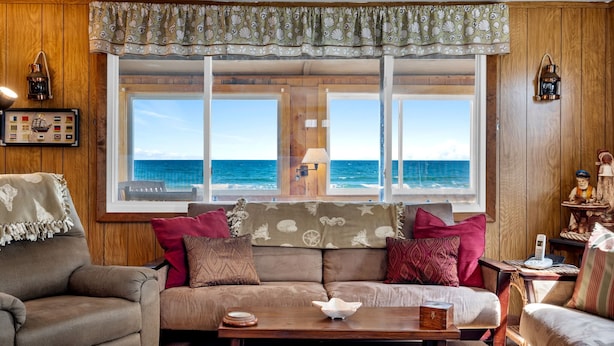 A cozy living room with a large window overlooking the sea, wooden walls, a sofa with patterned cushions, and a small table with decorative items. - Beachfront | 20 Steps to Long Sands | Ocean Views (York)