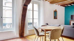 A room with a dining table and chairs, a sofa, and a large window with a view of the street. - Blueground | Ile Saint Louis, nr Notre Dame (Paris)