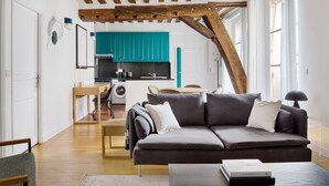 A modern living room with a grey sofa, wooden flooring, and a kitchen in the background with teal cabinets. - Blueground | Ile Saint Louis, nr Notre Dame (Paris)