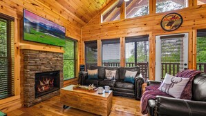A cozy cabin interior with a stone fireplace, leather sofas, and a large TV mounted on the wall.