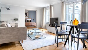 A modern living room with a sofa, a glass coffee table, a dining area with chairs, a TV, and a bed in the background. - Blueground | Invalides, nr Rodin & Maillol Museums (Paris)