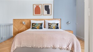 A bedroom with a wooden headboard, two pillows with paisley patterns, a beige blanket, and a nightstand with a lamp. - Blueground | Invalides, nr Rodin & Maillol Museums (Paris)