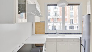A compact kitchen with white cabinets, a window with a view of buildings, and a hanging light fixture. - Blueground | Auteuil, elevator & concierge (Paris)