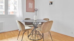 A modern dining area with a round glass table, four chairs, a white vase, and a wall-mounted light fixture. - Blueground | Auteuil, elevator & concierge (Paris)