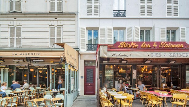 Deux cafés disposent d’espaces extérieurs aménagés avec des chaises et des tables en osier, et un bâtiment porte une enseigne indiquant « Restaurant ».