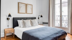 A bedroom with a bed, two bedside tables, a nightstand, a radiator, and a window with a view of a balcony. - Blueground | St Placide, balcony, nr Bon Marché (Paris)