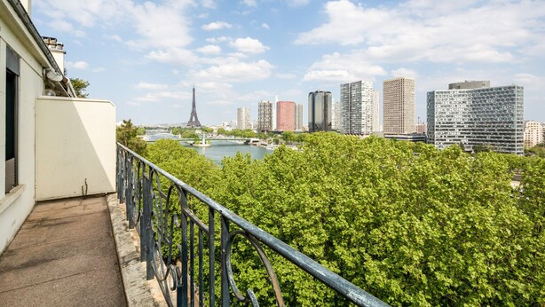 Vue sur la Tour Eiffel depuis un balcon doté d’une rambarde en métal, avec une rivière et des bâtiments modernes en contrebas.
