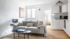 Un salon moderne avec un canapé gris, une table basse et un téléviseur. Il y a également un coin cuisine avec des meubles blancs et un four à micro-ondes.