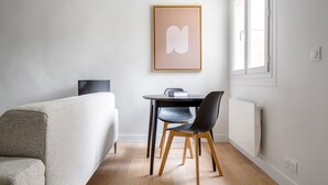 Une chambre moderne avec un lit, une petite table et une chaise, une œuvre d’art encadrée au mur et une fenêtre laissant entrer la lumière naturelle.