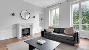 Un salón comedor amplio con chimenea, un sofá gris con cojines, una mesa de centro y grandes ventanales con vistas al verdor.