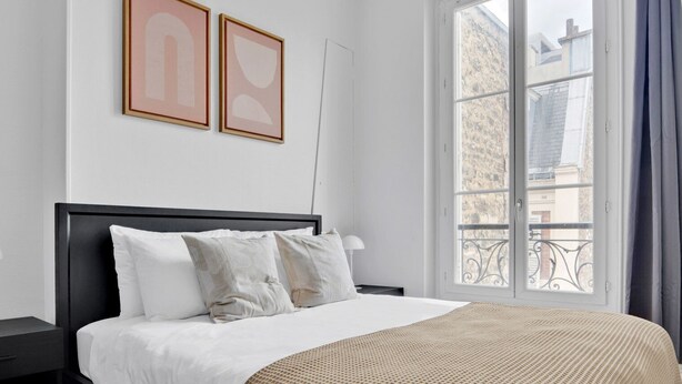 Una habitación de hotel con cama, mesita de noche, ventana con vista a edificios y dos cuadros en la pared.