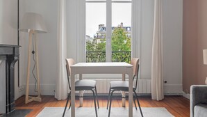A room with a dining table and chairs, a standing lamp, and a view of buildings through a large window. - Blueground | Arc de Triomphe, nr Parc Monceau (Paris)