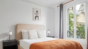 A neatly arranged bedroom with a large bed, two bedside tables with lamps, a framed picture on the wall, and a view of a building through a window with a curtain. - Blueground | Arc de Triomphe, nr Parc Monceau (Paris)
