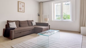 Ein modernes Wohnzimmer mit einem braunen Ecksofa, einem Glastisch, einer Lampe und einem Fenster mit Blick auf Grünflächen.