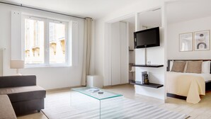 Ein modernes Hotelzimmer mit einer Couch, einem Glastisch, einem Bett mit beiger Decke, einem Wandfernseher und Kunstwerken an der Wand.