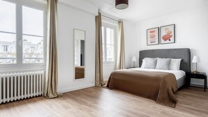 A modern bedroom with a large bed, a bedside table with lamps, a mirror, and a view of the city through large windows. - Blueground | Montparnasse, furnished & equipped (Paris)