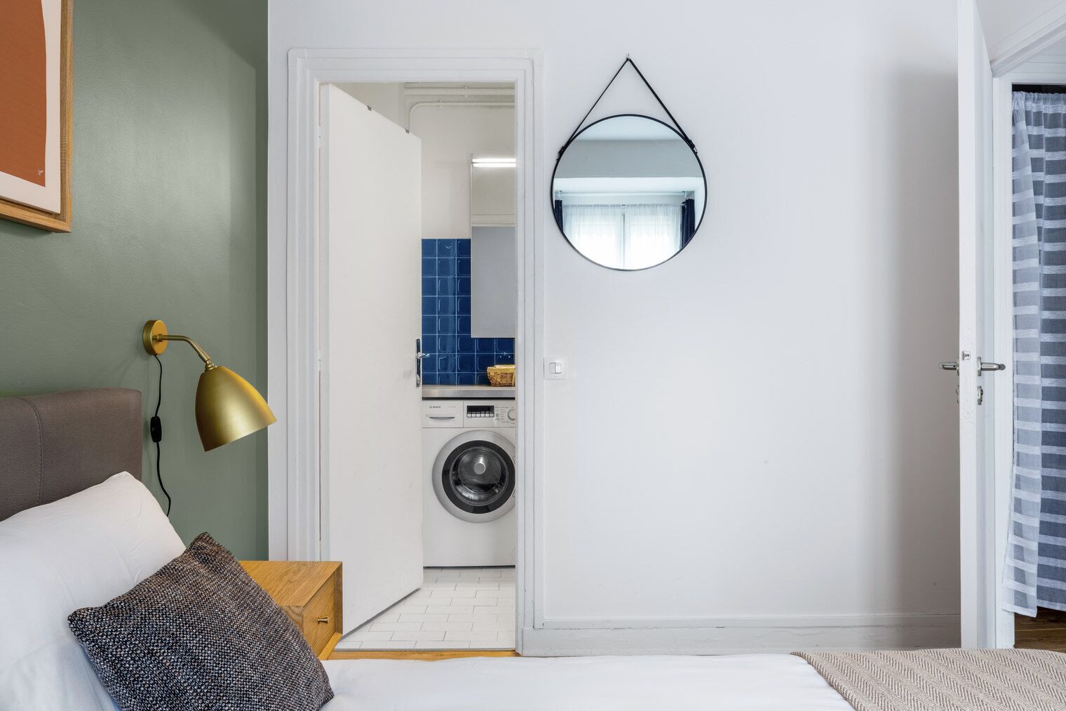 A bedroom with a bed, a bedside lamp, a mirror, and a view into a laundry room with a washing machine and blue tiles.