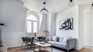 A modern living room with a grey sofa, a round coffee table, and a dining area with chairs and a table. There is a large window providing natural light and a painting on the wall. - Blueground | Montorgueil, concierge, furnished (Paris)