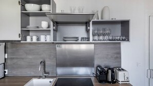 A modern kitchen with a stainless steel refrigerator, built-in oven, and a sink. There are shelves with cups, bowls, and wine glasses. - Blueground | Montorgueil, concierge, furnished (Paris)