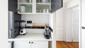 Une cuisine avec des meubles blancs, un plan de travail noir, un mixeur, une machine à café et un dosseret carrelé.