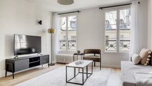 A modern living room with a flat-screen TV, a coffee table, a sofa, and two windows with balconies. - Blueground | Saint Lazare, elevator, near opera (Paris)