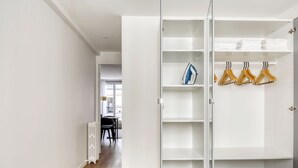 A white wardrobe with multiple shelves and hanging space, a radiator, and a view into a dining area with a table and chairs. - Blueground | Saint Lazare, elevator, near opera (Paris)