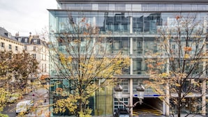 A modern building with a glass facade, a parking area with a few cars, and trees with autumn leaves in front.