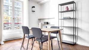 A modern dining area with a wooden table and four chairs, a black metal shelving unit with books, and a window with a view of buildings and trees.