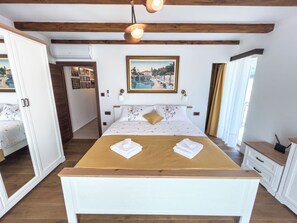 Een slaapkamer met een houten bedframe, wit beddengoed, een schilderij boven het bed en uitzicht op een gebouw door een raam.